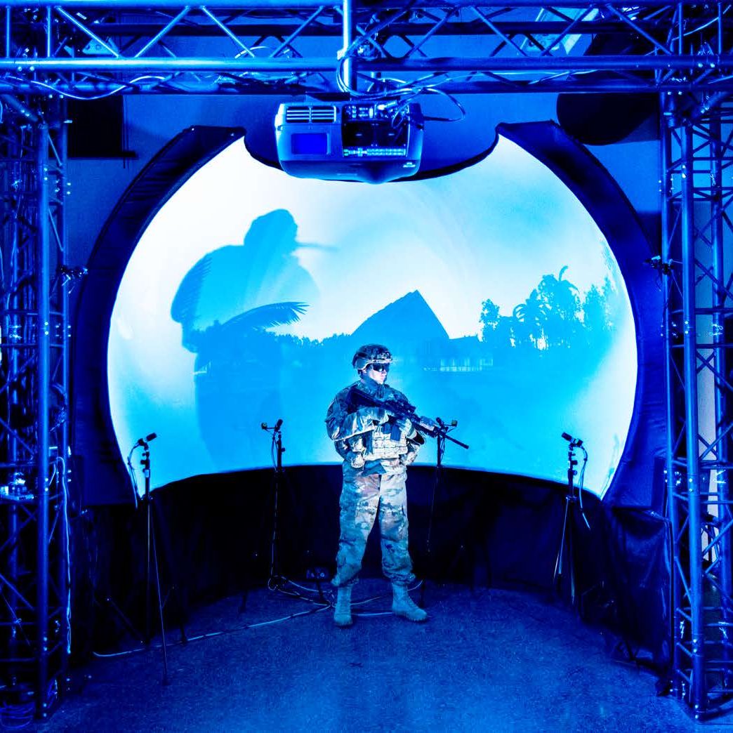 Warfighter holding a training weapon while standing in a simulation room surrounded by projectors and sensors. 