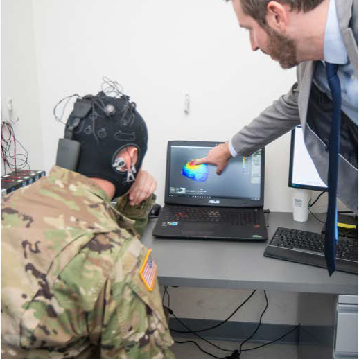Warfighter wearing sensors over the head and neck while sitting next to a researcher who explains the sensors and shows brain wave data on a laptop. 